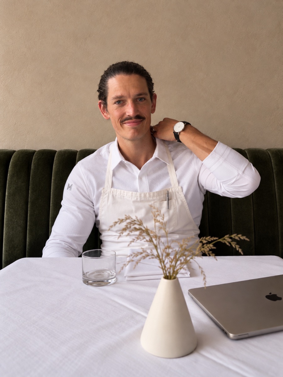 Greg seated in a velvet booth at a laid table, smiling, one hand at his collar